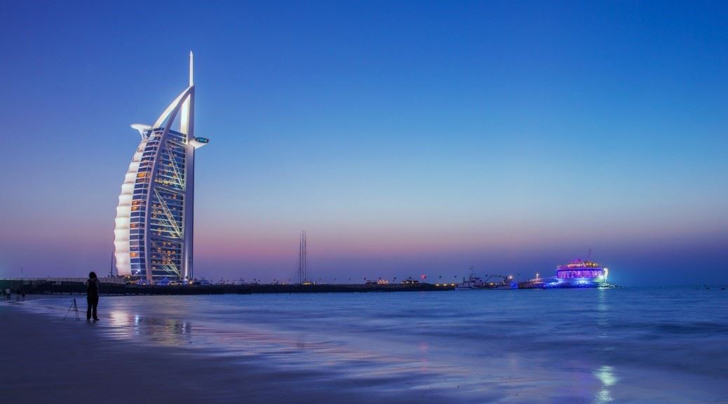 woman on jumeriah beach taking picture of burj al arab lit up with lights during night
